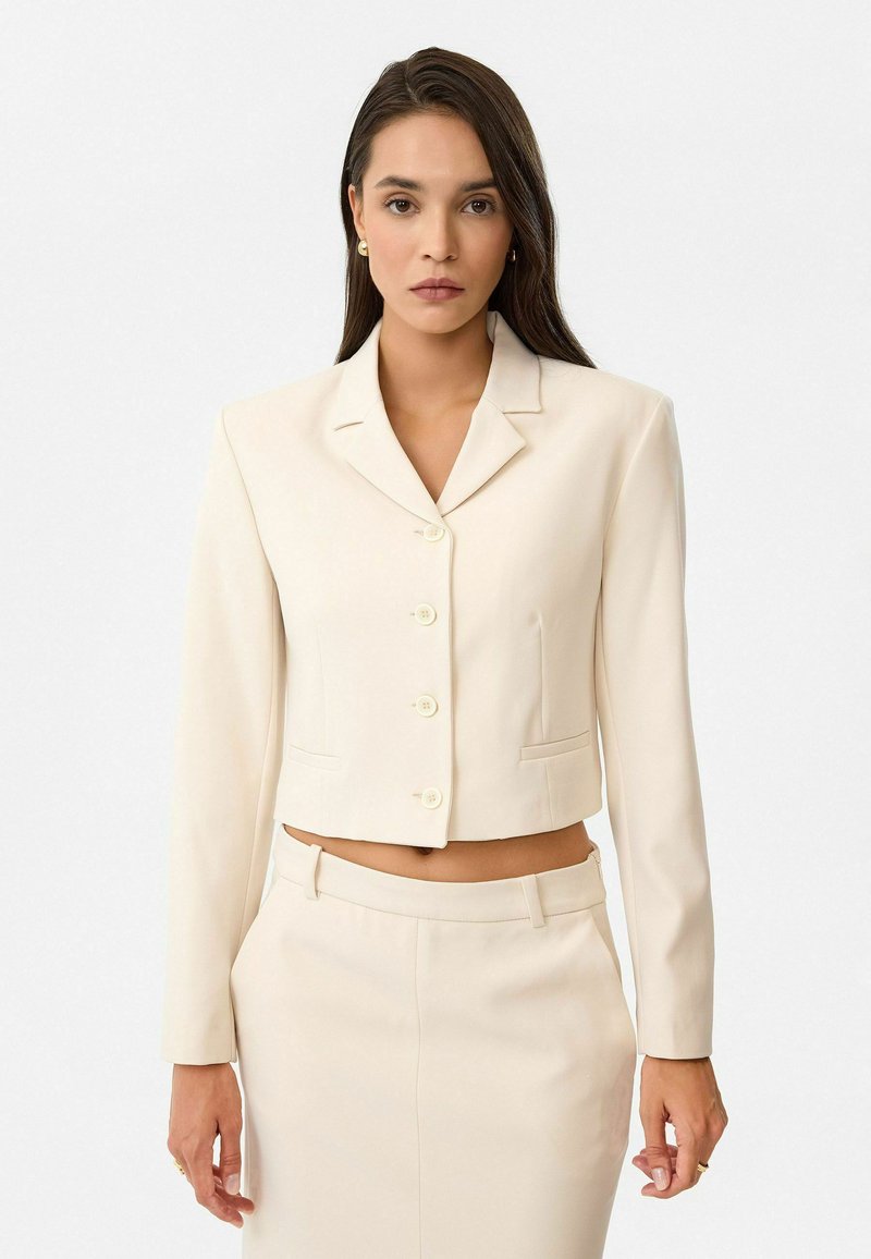 Blazer crème cintré avec un col, six boutons et des poches latérales, associé à une jupe crème assortie. Texture de tissu lisse.