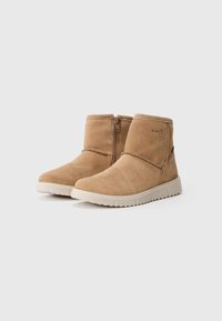 Une paire de bottines en suède beige avec des zips latéraux et des semelles en caoutchouc blanc sur un fond blanc uni.