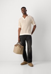 Beige gestrickter Poloshirt mit V-Ausschnitt und kurzen Ärmeln, kombiniert mit dunklen Jeans und einer tanfarbenen Lederhandtasche. Die Schuhe haben ein klobiges, legères Design.