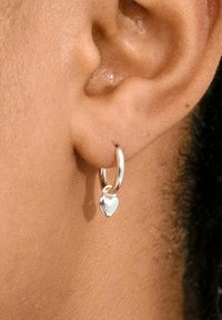 Silvrig hoopörhänge med en liten hjärtcharm, slät yta, cirkelformad design och glänsande finish, bärs på kanten av ett öra.