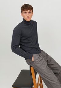 Pull à col roulé gris foncé en tissu doux, associé à un pantalon rayé gris clair, porté par un modèle assis.