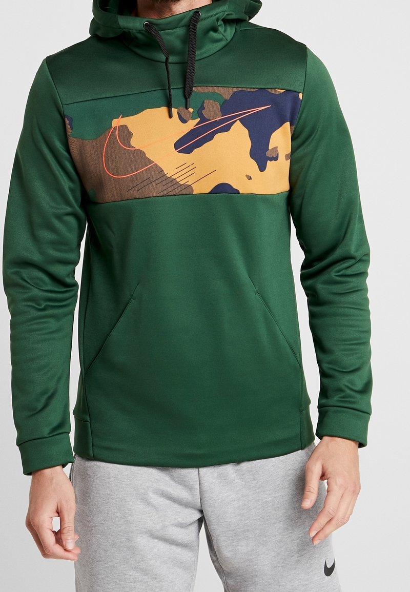 Homme portant un sweat à capuche vert foncé avec un panneau camouflage multicolore sur la poitrine et un logo Nike orange, associé à un short Nike gris clair.