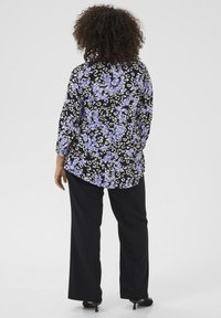 Schwarze Bluse mit lila und weißen Blumenmuster, lange Ärmel, lockere Passform. Kombiniert mit schwarzer weit geschnittener Hose und Absatzschuhen.