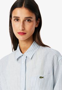 Camisa de botones a rayas en azul claro y blanco, hecha de algodón, con un pequeño logotipo de cocodrilo bordado en verde en el bolsillo del pecho izquierdo.
