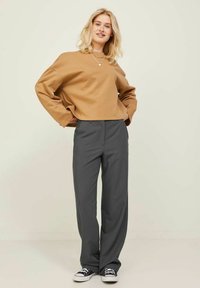 Korte sweatshirt van lichtbruin stof, gecombineerd met losse grijze broeken. Heeft brede mouwen en zwarte sneakers. Soepele texturen overal.