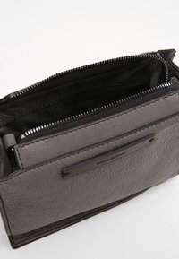 Bolso de cuero gris con superficie texturizada, cierre de cremallera, compartimento interior y bolsillo delantero con parche de logo.