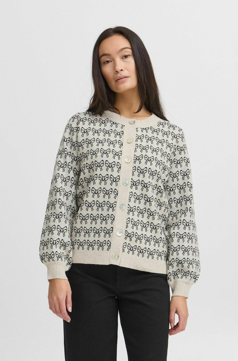 Cardigan tricoté beige avec un motif de nœud noir, doté d'un col rond, de longues manches bouffantes et d'une fermeture à boutons à l'avant.