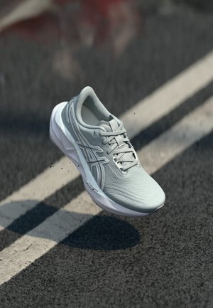 Lys grå løpesko med hvit såle i luften over asfaltvei med hvite kjørefeltsmarkeringer.