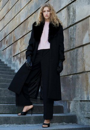 Femme aux cheveux bouclés se tient sur des marches en pierre, portant un manteau noir avec un col en fourrure, des gants noirs, un pantalon noir, un haut rose et des sandales à talons noires.