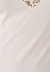 Blusa beige chiaro con scollo a V, caratterizzata da un delicato inserto in pizzo e una texture a coste, tessuto liscio e dettagli di cucitura sottili.