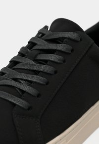 Gros plan d'une basket noire avec des lacets noirs texturés et une semelle blanche, montrant des coutures détaillées et des œillets sur la partie supérieure de la chaussure.