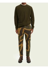 Persoon draagt een donkergroene trui, bruine en groene camouflage cargobroek, en donkergroene laarzen, staand tegen een lichte achtergrond.