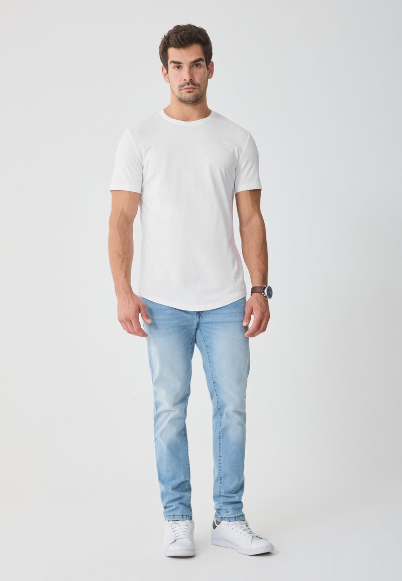 Camiseta blanca de cuello redondo y mangas cortas combinada con vaqueros de mezclilla azul claro. Usada con zapatillas blancas, presenta un reloj de cuero en la muñeca izquierda.