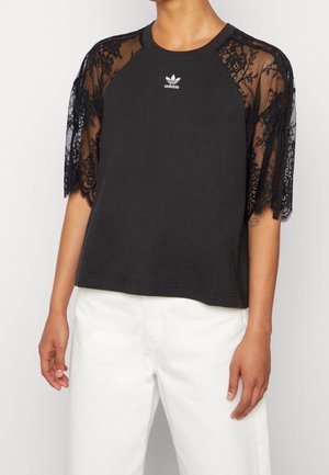 T-shirt noir Adidas avec manches courtes en dentelle florale, col rond et logo blanc sur la poitrine, porté avec un pantalon blanc.