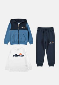 Sweat à capuche bleu et marine avec fermeture éclair, pantalon à cordon de serrage marine, et chemise blanche à manches longues, tous avec le logo ellesse, disposés à plat sur un fond blanc.