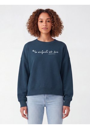 Frau mit lockigem Haar, die einen dunkelblauen Pullover mit dem deutschen Text "So einfach ist das" und hellblaue Jeans trägt, vor weißem Hintergrund stehend.