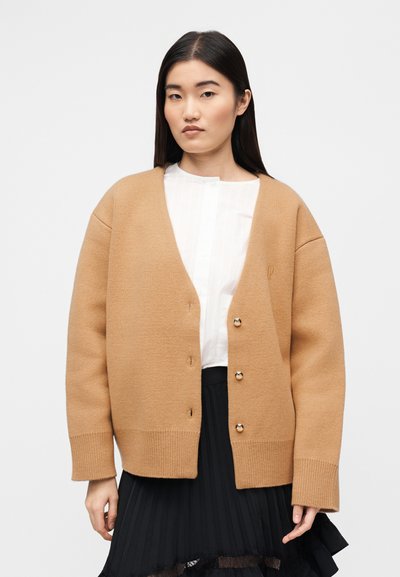 Μπεζ ζακέτα με λαιμόκοψη V, κουμπιά μπροστά και ριμπ μανσέτες. Διαθέτει απαλό ύφασμα και φοριέται πάνω από ένα λευκό πουκάμισο με γιακά και μαύρη φούστα.