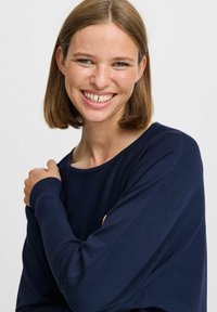 Pull à manches longues bleu marine en maille côtelée ; col rond et coupe décontractée. Le modèle pose avec les bras croisés, souriant.