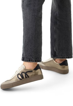 Baskets beiges avec des accents noirs, présentant une tige en cuir lisse, un bout rond et une semelle en caoutchouc texturé. Les lacets sont de couleur crème.