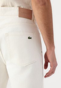 Balti džinsai su ruduo odiniais Lacoste etiketėmis ant liemens ir žaliai siuvinėtų krokodilo logotipu ant galinės kišenės.