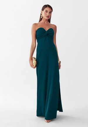 Femme en robe bustier bleu sarcelle foncé avec corsage torsadé, tenant une pochette dorée et portant des boucles d’oreilles et bracelets dorés, debout devant un fond uni.