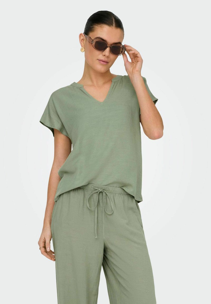 Olivgrön tvådelad outfit som har en V-ringad topp med korta ärmar och byxor med dragsko. Materialet verkar lätt och har en slät textur.