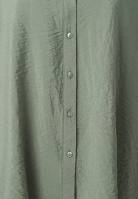 Camisa verde claro de botões, feita de tecido enrugado, com seis botões redondos e uma superfície texturizada, apresentando costura visível ao longo da pala.