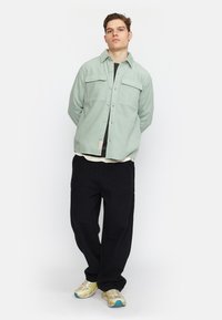 Lichtgroen, knoopshirt met twee voorkruizen, gedragen over een zwart shirt; zwarte broek en neutraalgekleurde sneakers maken de outfit compleet.