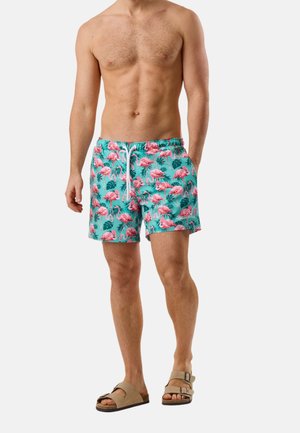 Mand iført turkise badeshorts med pink flamingo-mønster, beige sandaler, stående med den ene hånd i lommen mod hvid baggrund.