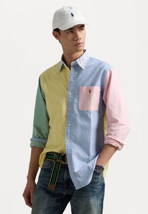 Farbblockiertes Button-Up-Hemd mit gelben, grünen, blauen und pinken Abschnitten, einer Brusttasche und gesticktem Logo. Kombiniert mit Jeans und einer Mütze.