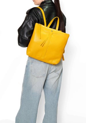 Donna con giacca di pelle nera e jeans azzurri porta uno zaino grande in pelle gialla sulla schiena.