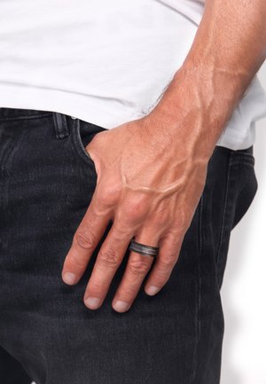 Black metal ring with a textured surface, worn on a hand resting in a pocket. The hand has visible veins and is positioned against dark jeans.
