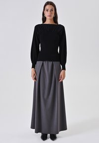 Donna che indossa una maglietta nera a maniche lunghe e una gonna maxi grigia a vita alta, in piedi con le mani rilassate ai lati.