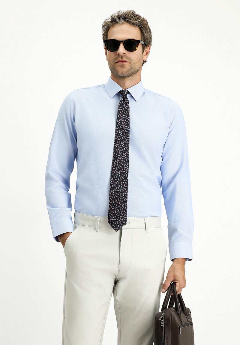 Camicetta blu chiaro, cravatta a motivi floreali, pantaloni beige chiaro, tiene una borsa di pelle marrone, indossa occhiali da sole neri.