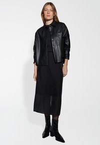 Veste chemise en cuir noir avec un bouton à l'avant, associée à une longue jupe noire transparente et des bottines noires brillantes. Design minimaliste.