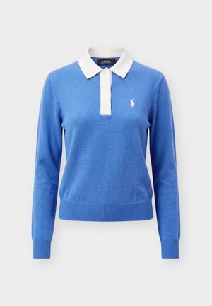 Personne portant un polo bleu manches longues avec un col blanc, un pantalon bleu marine foncé, une ceinture beige et des baskets blanches, debout devant un fond uni.
