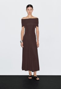 Hnědá maxi šaty se širokými ramínky, které mají fitted korpus a splývavou sukně. Hladká, pružná látka a minimalistický design bez vzorů.