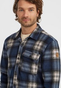 Groot geruit flanellen shirt in de kleuren blauw, zwart en wit, met een borstzak, drukknopen en een ronde logo-patch op de mouw.