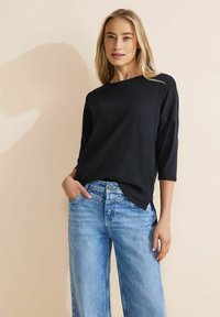 Schwarzes, strukturiertes Oberteil mit Dreiviertelärmeln, kombiniert mit hellblauen, weiten Jeans. Das Oberteil hat eine lockere Passform und seitliche Schlitze.