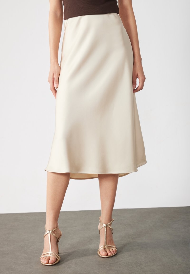 Jupe midi en satin beige avec un tombé fluide et une fente latérale, associée à des sandales métalliques à brides. Silhouette simple, texture lisse.