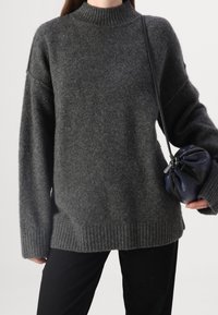 Donkergrijze, oversized gebreide trui met een hoge kraag en ribbelzoom. Zachte textuur, gedragen met een kleine, geplooide handtas in marineblauw en zwart.