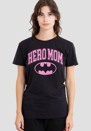 Woman with brown hair wearing a black t-shirt with pink "HERO MOM" text and a pink Batman logo on a plain gray background.