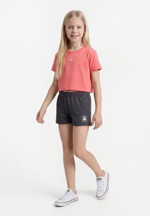 Jeune fille debout portant un t-shirt court corail, un short gris foncé avec logo, et des baskets blanches sur fond blanc uni.