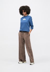 Μπ λε φούτερ με λευκό λογότυπο, συνδυασμένο με καφέ παντελόνι από βελούδο με ρυθμιζόμενο κορδόνι στη μέση. Τονισμένο με ανοιχτόχρωμα αθλητικά παπούτσια και γυαλιά ηλίου.