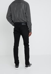Hombre con pantalones vaqueros ajustados negros, camisa blanca y negra a rayas verticales, y zapatillas grises, de pie de espaldas frente a un fondo liso.