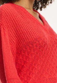 Maglione rosso lavorato a maglia con scollo a V, caratterizzato da un motivo testurizzato e maniche ampie. Il tessuto è morbido e leggero, aumentando il suo fascino casual.
