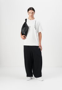 Hvid t-shirt med et grå logo, parret med sorte vide bukser og hvide sneakers. En sort sling taske bæres på tværs af kroppen.