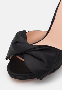 Melnas augstpapēžu kurpes ar maigu satīna loku priekšpusē, slēgtas pirkstgalos un gludu, kontrastējošu beigu iekšpusi.