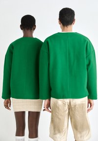 To personer har på seg overdimensjonerte grønne topper laget av et teksturert stoff. Den ene kombinerer det med en lys beige skjørt, mens den andre har på seg skinnende beige shorts.