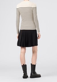 Personne portant un haut à manches longues rayé crème et noir, une jupe plissée noire et des bottes noires mi-mollet, se tenant sur un fond gris clair.